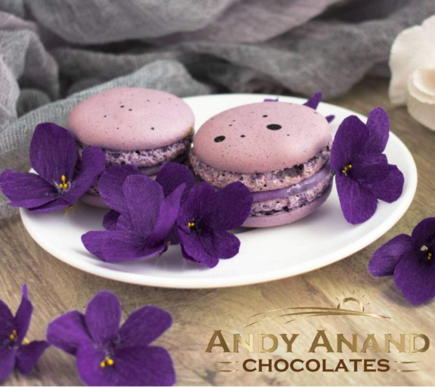 Sweet and colorful French macarons presented on a plate with fresh purple flowers, showcasing the delicious and visually appealing nature of this product. Andy Anand Chocolates, All Products,Products,Pies,Fresh Baked Macaron