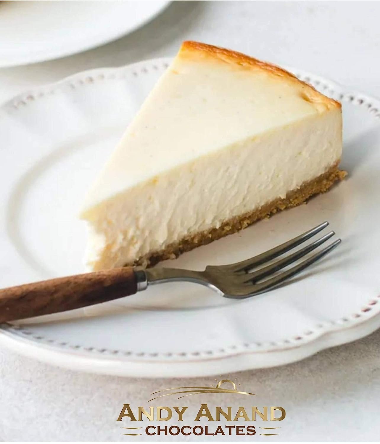 Slice of creamy New York style cheesecake on a white plate with a fork, handcrafted and freshly baked by Andy Anand Chocolates.