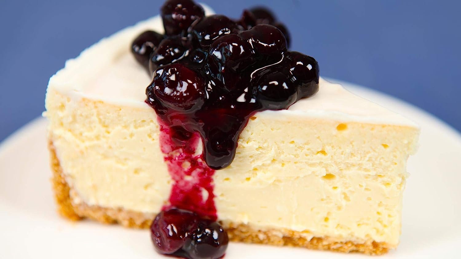 Slice of New York style cheesecake with blueberry topping on white plate against blue background