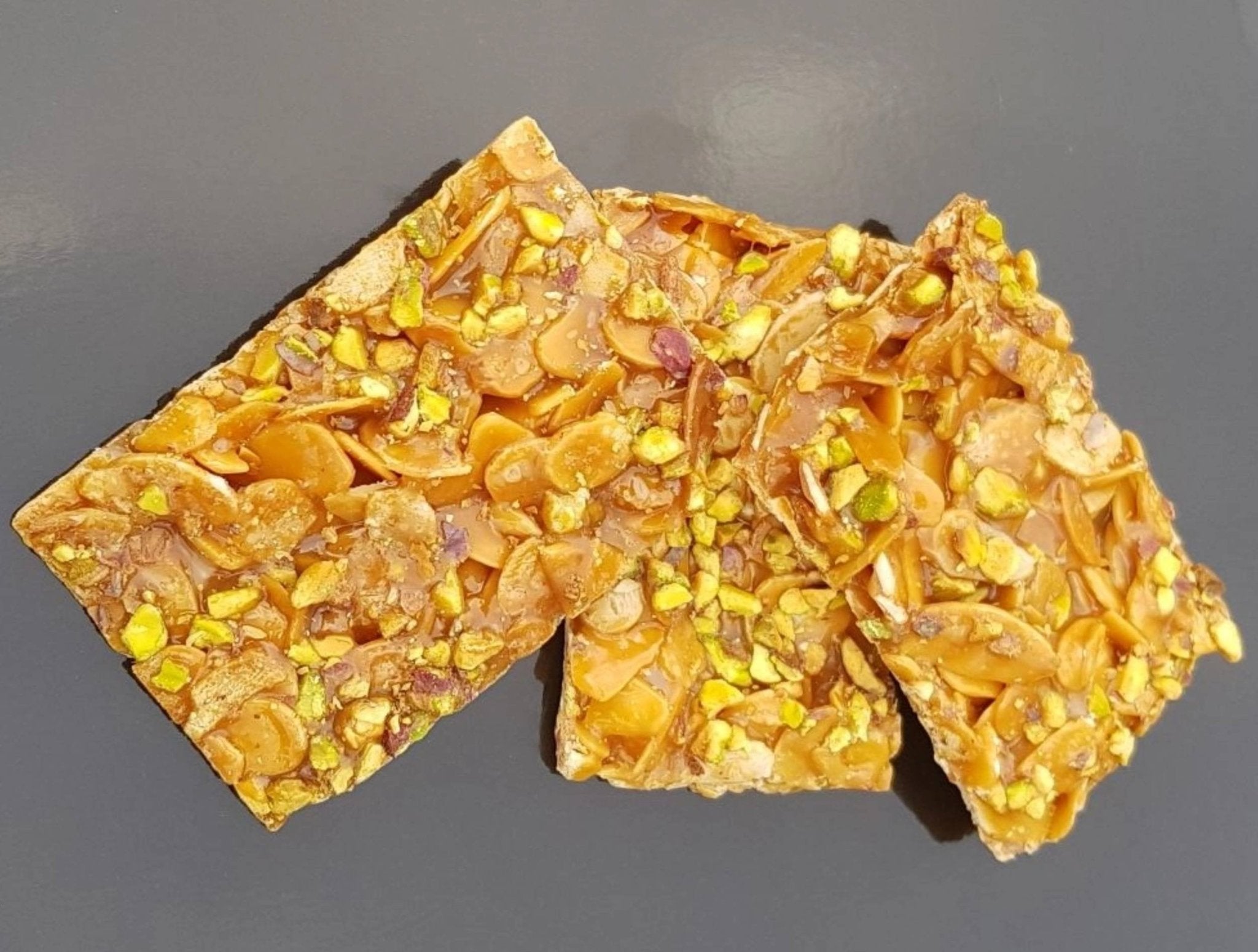 Three pieces of crunchy Italian pistachio brittle with sliced almonds and pistachio nuts on a dark background