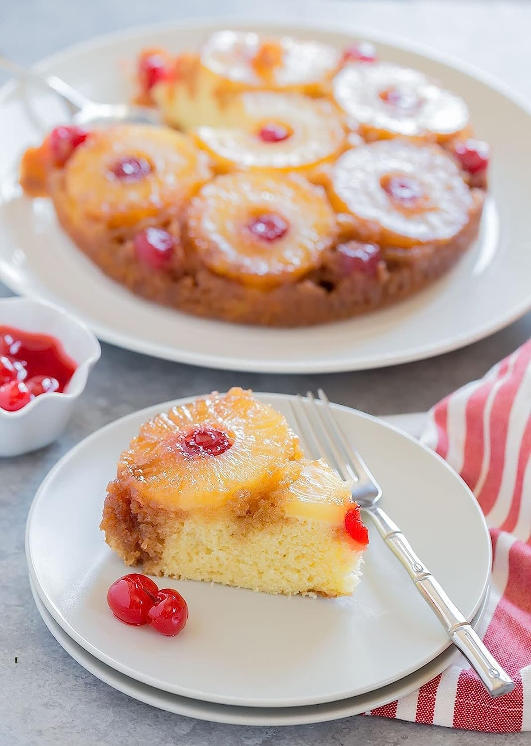 Andy Anand Traditional Pineapple Upside Down Cake with glazed pineapple rings and cherries, slice served on a white plate with a fork.