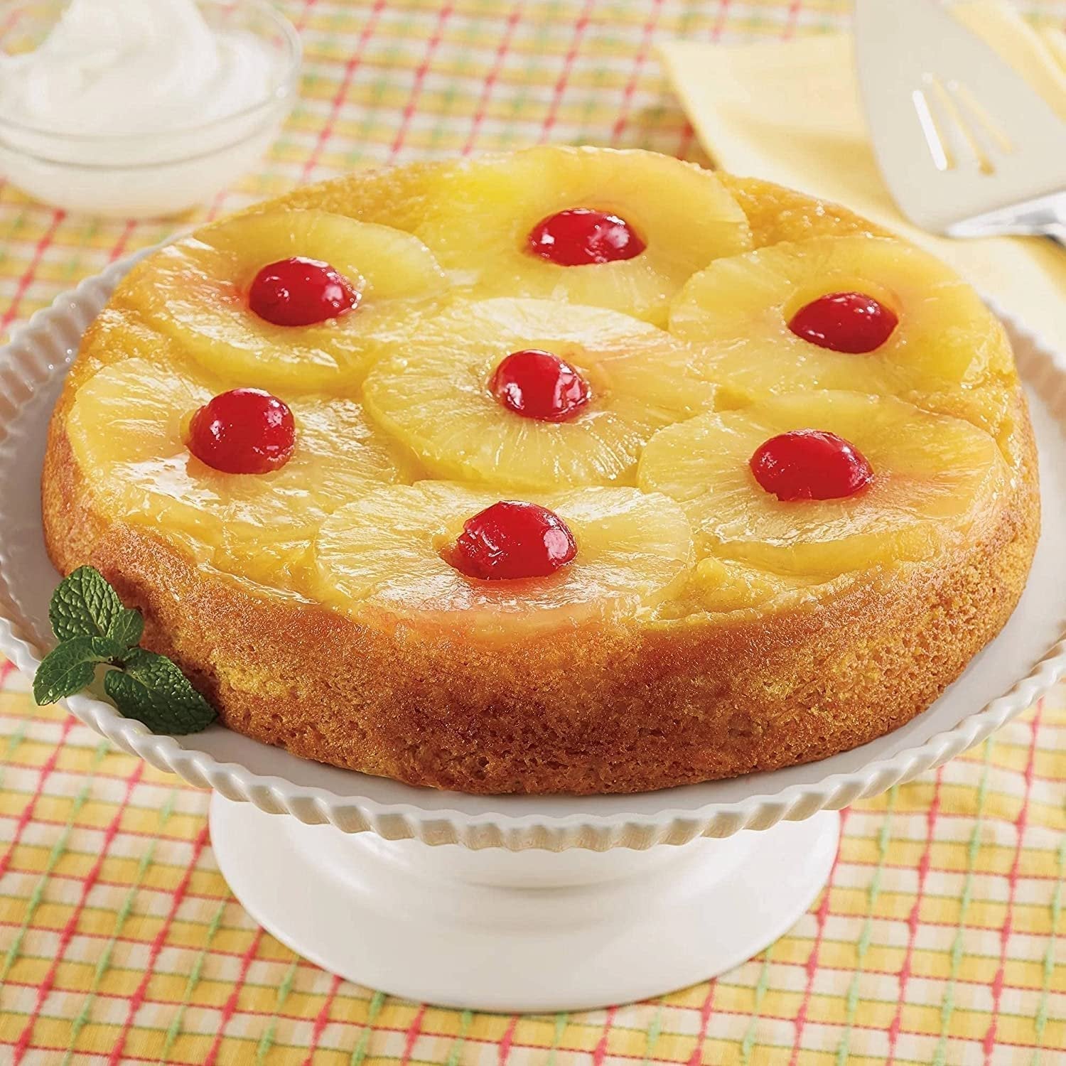 Andy Anand Traditional Pineapple Upside Down Cake with pineapple rings and cherries on a white cake stand
